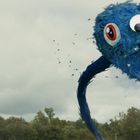 Ein großes blaues Wesen mit einem großen Auge erhebt sich über Bäume unter einem bewölkten Himmel.