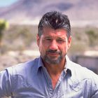 Ein Mann mit Bart und kurzen Haaren trägt ein helles Hemd. Im Hintergrund ist eine Wüstenlandschaft zu sehen.