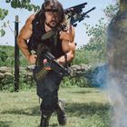 Ein muskulöser Mann rennt mit einer großen Waffe über eine Wiese, umgeben von Bäumen und Steinen, Rauch im Vordergrund.