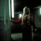 Keine erkennbaren Texte. Ein Mädchen in rotem Mantel berührt eine Lichtsäule in einer dunklen Umgebung.