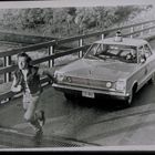 Ein Mann rennt auf einer Brücke. Ein Polizeiauto folgt ihm dicht, während ein Polizist im Wagen sitzt.