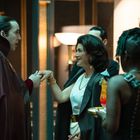 Ein Mann im langen Umhang küsst die Hand einer elegant gekleideten Frau; eine Kellnerin serviert Cocktails.