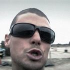 Eine Person mit Sonnenbrille im Vordergrund öffnet den Mund. Im Hintergrund steht ein blaues Auto in einer offenen Landschaft.