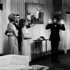 Im Raum steht ein Mann mit erhobenen Händen, während drei andere Personen auf ihn zeigen und miteinander interagieren.