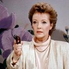 Es gibt keinen erkennbaren Text. Eine Frau mit kurzem Haar hält ein Objekt. Im Hintergrund sind lilafarbene Blumen.