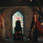 Zwei Frauen stehen gegenüber in einem mittelalterlich aussehenden Raum mit buntem Glasfenster und Rüstungen.