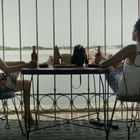 Zwei Frauen sitzen an einem Tisch mit Getränken, vor einem Geländer mit Blick auf Wasser. Im Hintergrund ist ein Hafen zu sehen.