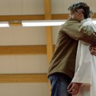 Zwei Personen umarmen sich in einer Umgebung mit Holzbalken an der Decke. Eine trägt eine braune Jacke, die andere Weiß.