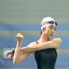 Eine Schwimmerin mit Badehaube und Schwimmbrille dehnt ihren Arm in einer Sporthalle.