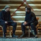 Zwei Männer in schwarzer Kleidung sitzen gegenüber auf Stühlen vor einem geschlossenen Rolltor mit rotem X.