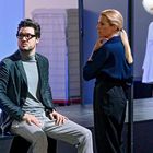 Ein Mann und eine Frau in eleganter Kleidung stehen und sitzen, im Hintergrund läuft eine Person in weißem Bademantel.