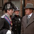 Ein Mann in Uniform mit Orden spricht mit einem eleganten Herrn in Mantel und Hut, beide neben einem Polizisten.