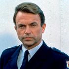 Ein Mann in einer dunkelblauen Uniform mit Abzeichen, ernster Gesichtsausdruck, kurz geschnittenes, dunkles Haar.