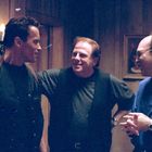 Eine warm beleuchtete Szene mit drei Männern, die sich unterhalten und lachen; die Stimmung ist entspannt und freundlich.