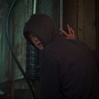 Neonschrift "NLS". Person mit Kapuze schaut über die Schulter in einem dunklen, urbanen Hinterhof.
