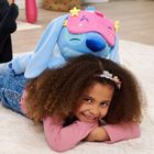 Ein glückliches Kind liegt lächelnd auf dem Boden. Ein blaues Plüschtier mit rosa Schlafmaske ruht auf seinem Rücken.