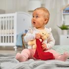 Ein Baby sitzt auf einer Decke, hält ein Stofftier und schaut interessiert in die Ferne, im Hintergrund ein Kinderbett.