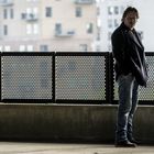 Eine Person in Jeans und Mantel steht seitlich an einem gelochten Geländer vor verschwommener Stadtansicht.