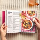 Text: "Cheat's Bouillabaisse (Classic French Fish Soup)". Rezeptbuch mit Suppe und Brot. Glas Tee daneben. Person hält das Buch.