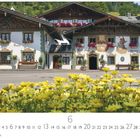 "Bayrischer Dorfplatz"; Häuser mit bemalten Fassaden, buntblühende Blumen in Kästen und im Vordergrund gelbe Blüten.