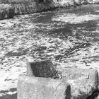 Betonblock im Wasser vor einer grasigen Böschung, umgeben von Schaum auf der Wasseroberfläche.