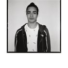 Ein junger Mann mit zusammengebundenem Haar trägt ein Shirt mit Vogelmuster und eine Jacke mit Streifen.