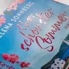 "Unser schönster Sommer" von Elena Sonnberg ist ein Roman. Pastellfarbener Hintergrund mit Blumen und einem Segelboot.