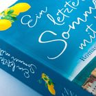 Text: "Ein letzter Sommer mit dir", "Roman". Hellblaues Cover mit Zitronen und Küstenlandschaft.