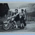 Kennzeichen: 0.78.541. Zwei Frauen sitzen lachend auf einem Motorrad vor einem Haus mit Bergen im Hintergrund.