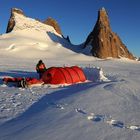 Keine erkennbaren Texte. Eine Person steht bei einem roten Zelt im Schnee, dahinter ragen imposante Berge empor.