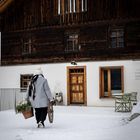 „HANSL HOF“, eine Person mit Korb geht durch Schnee zu einem Holzhaus.