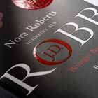 Text: "Nora Roberts, schreibt als J.D. Robb, Blutiges Band, Roman, blanvalet." Buchcover in dunklen Tönen mit rotem Akzent.