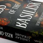 Text: "KISS OF THE BASILISK", "VERFÜHRERISCHES GIFT", "blush". Dunkler Hintergrund mit roten Blumen.