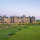Ein großes Steingebäude im Herrenhausstil, umgeben von einem gepflegten Golfplatz, unter klarem Himmel.