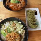 Zwei Schalen mit Nudelsuppe und Gemüse, eine mit frittiertem Gericht. Daneben sechs grüne Teigtaschen mit Sojasauce.