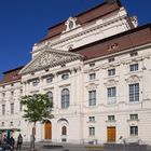 Großes, historisches Gebäude mit verziertem Giebel und mehreren großen Fenstern, zwei Holztüren, Bäume davor.