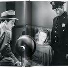 Ein Mann in Anzug und Hut spricht mit einer Frau, die von einer Lampe beleuchtet wird; daneben steht ein Polizist.