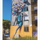 06, Kleines Gebäude mit Wandgraffiti einer nachdenklichen Figur mit buntem Afro, blauer Himmel im Hintergrund.