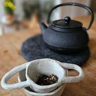 Graue Teetasse mit losem Tee darin auf einem Holztisch, dahinter eine schwarze Teekanne.