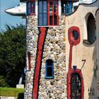 6 2027. Ein Turm mit Steinfassade, bunten Fenstern, rotem Mosaik und einer goldenen Kuppel. Flugzeug im Hintergrund.