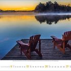 6 2023. Zwei Holzstühle auf einem Steg mit Blick auf einen ruhigen See bei Sonnenuntergang, leichte Nebelschwaden über dem Wasser.