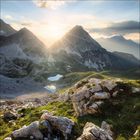Eine felsige Berglandschaft bei Sonnenuntergang, mit grasbewachsenen Flächen und einem kleinen See in der Ferne.