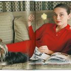 Eine Frau in roter Kleidung liegt auf einem Sofa, hält eine Blume und hält ein geöffnetes Buch in der Hand. Ein Hund liegt neben ihr.