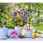 06, Kalender Juni. Blumenarrangement auf Tisch mit weißem Geschirr, Vase mit gelben und lila Blumen, grüne Bäume im Hintergrund.