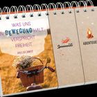 Text: "WAS UNS IN BEWEGUNG HÄLT, VERSPRICHT FREIHEIT. ANGELIKA EMMERT." Daneben Illustrationen eines Krabbenlogos und eines Lagerfeuers.