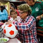 Eine Frau unterschreibt einen rot-weißen Fußball in einem belebten Raum mit mehreren Personen um einen Tisch.