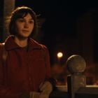 Eine Frau in einem roten Mantel steht im Freien bei Nacht und blickt leicht lächelnd in die Ferne. Im Hintergrund Lichter.