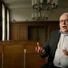 Ein Mann gestikuliert in einem leeren Saal mit Holzbänken und Kronleuchter. Er trägt Anzug und Hemd.