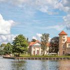 Text: "DER NORDWESTEN". Ufer mit Bäumen und Gebäuden reflektiert im Wasser, blauer Himmel mit Wolken.