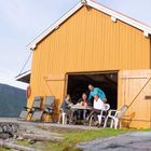 Vier Personen sitzen an einem Tisch vor einem gelben Holzhaus in einer ländlichen, grünen Umgebung.